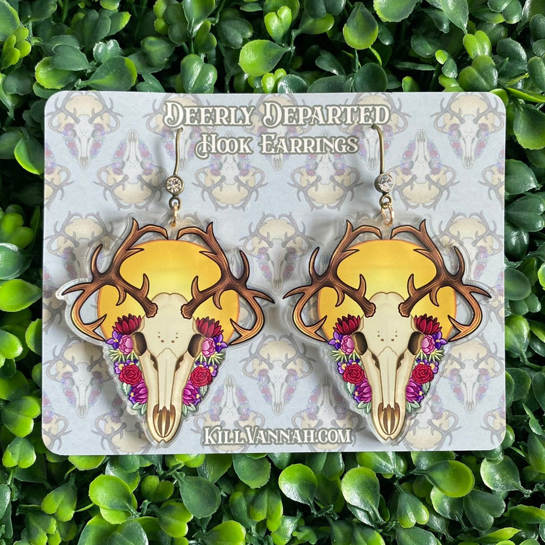 Earrings with bronze hooks and a clear rhinestone, featuring a deer skull in front of a yellow sun, surrounded by red, pink, purple and green flowers on a cardstock background featuring a pattern of the same design. Both are lying on a fake leaves tile..
