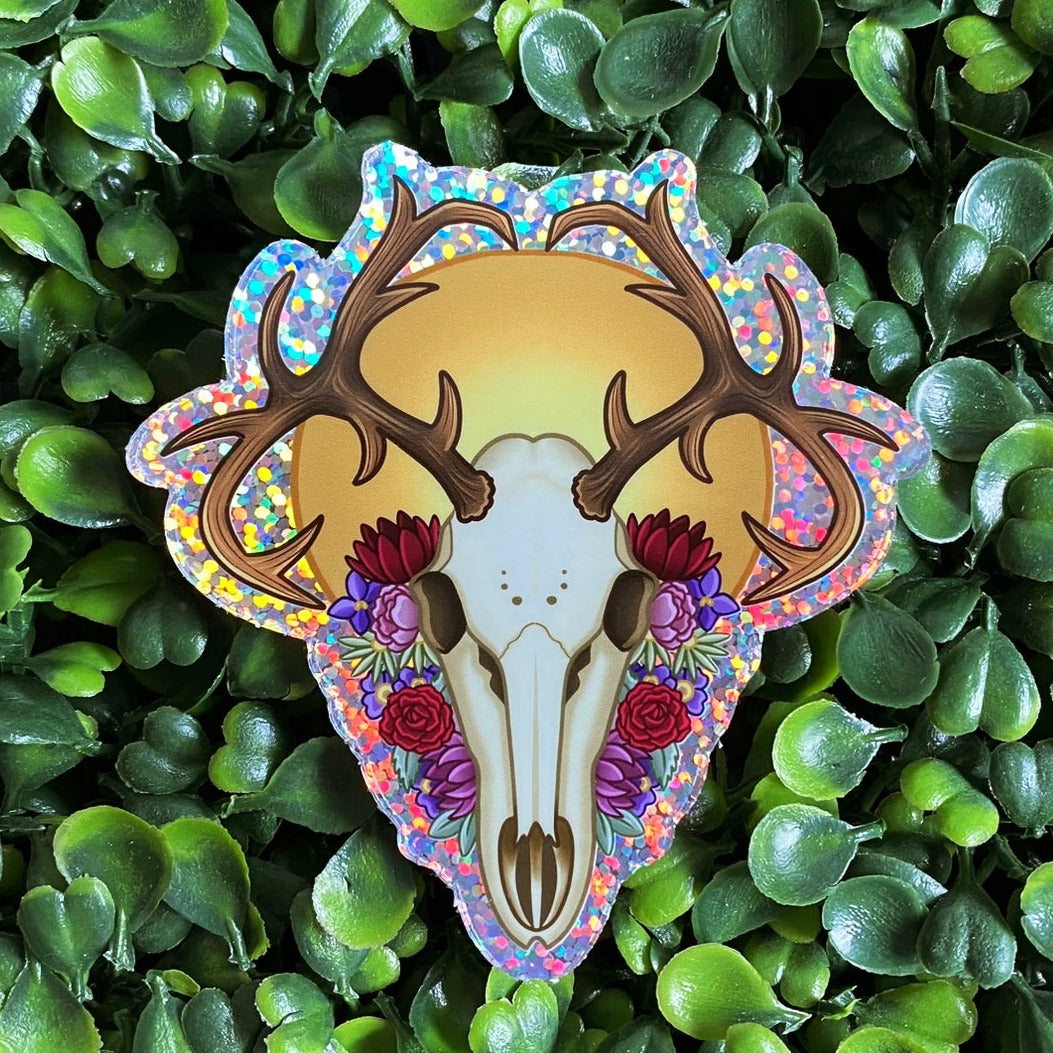 Glitter holographic sticker featuring a deer skull with a sun behind it and an assortment of flowers in red, pink, purple and green.
