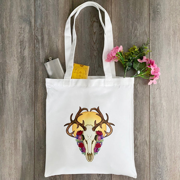 White tote bag lying on a wooden floor backdrop. The tote bag features a design of a deer skull in front of a yellow sun, surrounded by flowers in red, pink, purple, and green. There's a silver flask, a deck of cards & flowers in the bag.