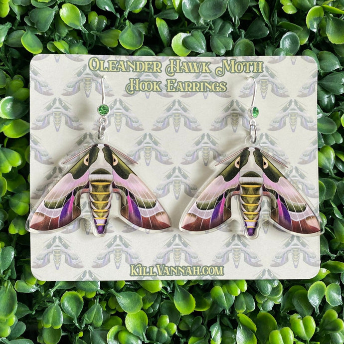 Hook earrings with a silverplated hook and green rhinestone, with an illustration of an Oleander Hawk Moth attached to it on clear acrylic. On a background of cardstock featuring the illustration as a pattern.